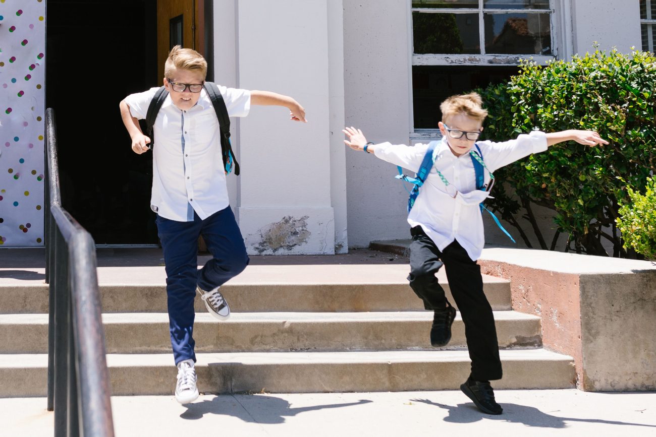 Two boys with backpacks energetically jumping down concrete steps outside a school.